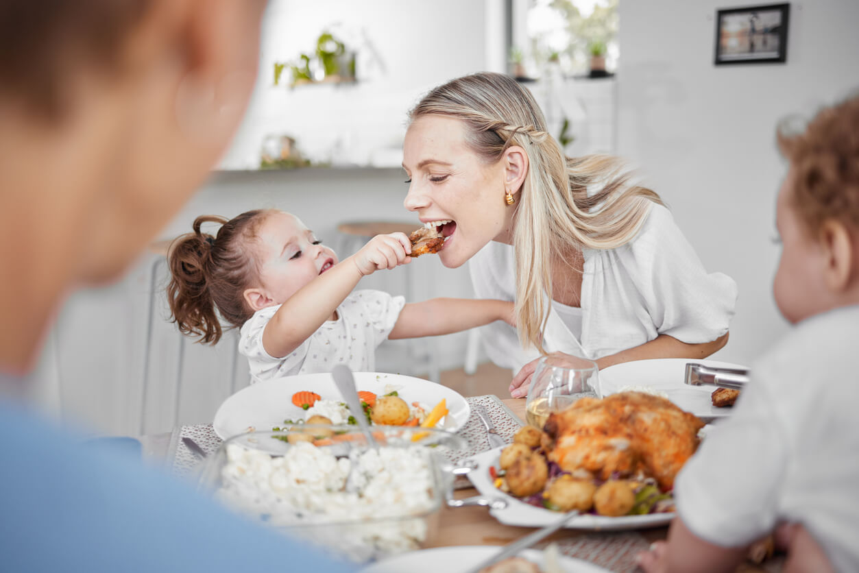 At the dining table, a mum plays with her young daughter, pretending to take a bite of her food.