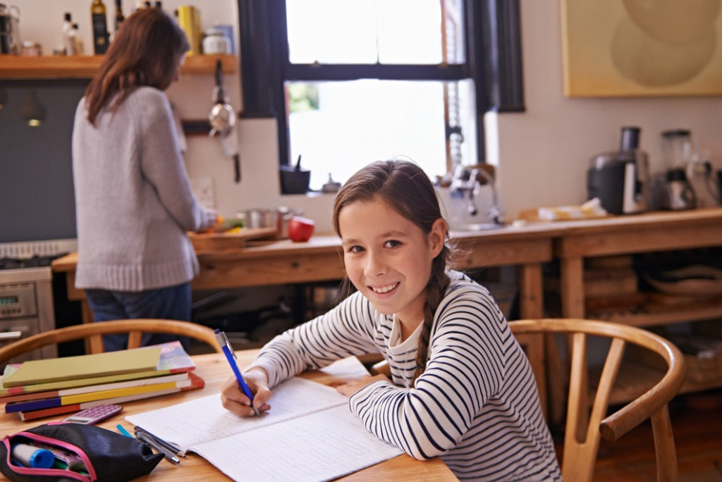 A young girl sits at a dining table doing her homework while her mum prepares a meal